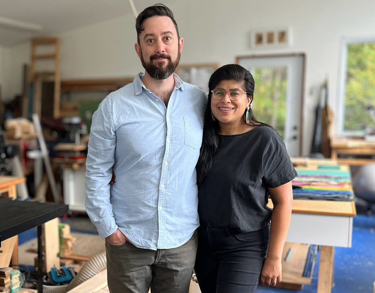 A man and a woman standing together in a workshop or studio space with tools, wood, and tables visible in the background.